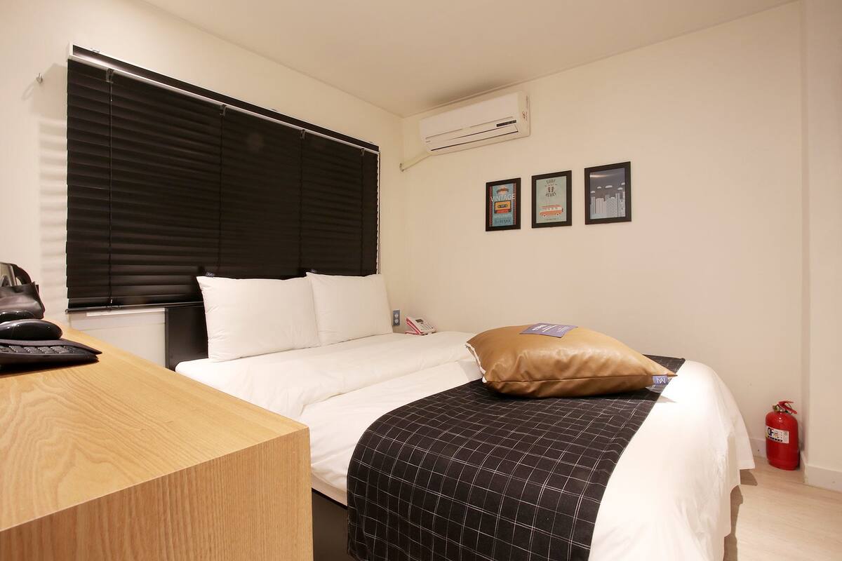 A hotel room with a bed, a desk, a telephone, a fire extinguisher, and wall-mounted air conditioning.