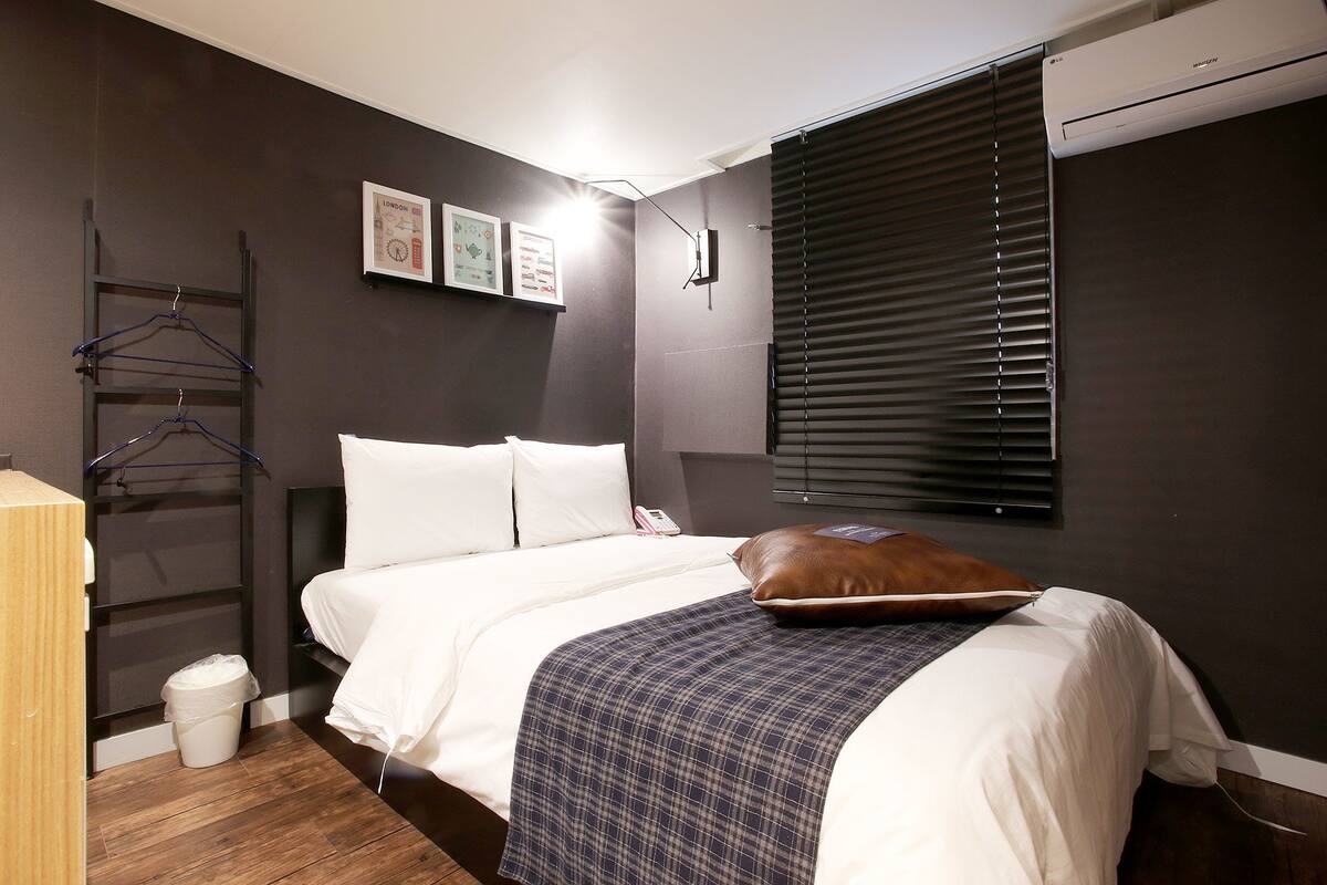 A bedroom with a bed, a bedside table, a hanging clothes rack, a wall-mounted air conditioner, and a window with blinds.