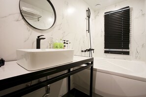 A bathroom with a white sink, black faucet, and a black-framed mirror. There are toiletries on the sink, a showerhead, and a window with blinds. - Kotel Sinchon (Seoul)