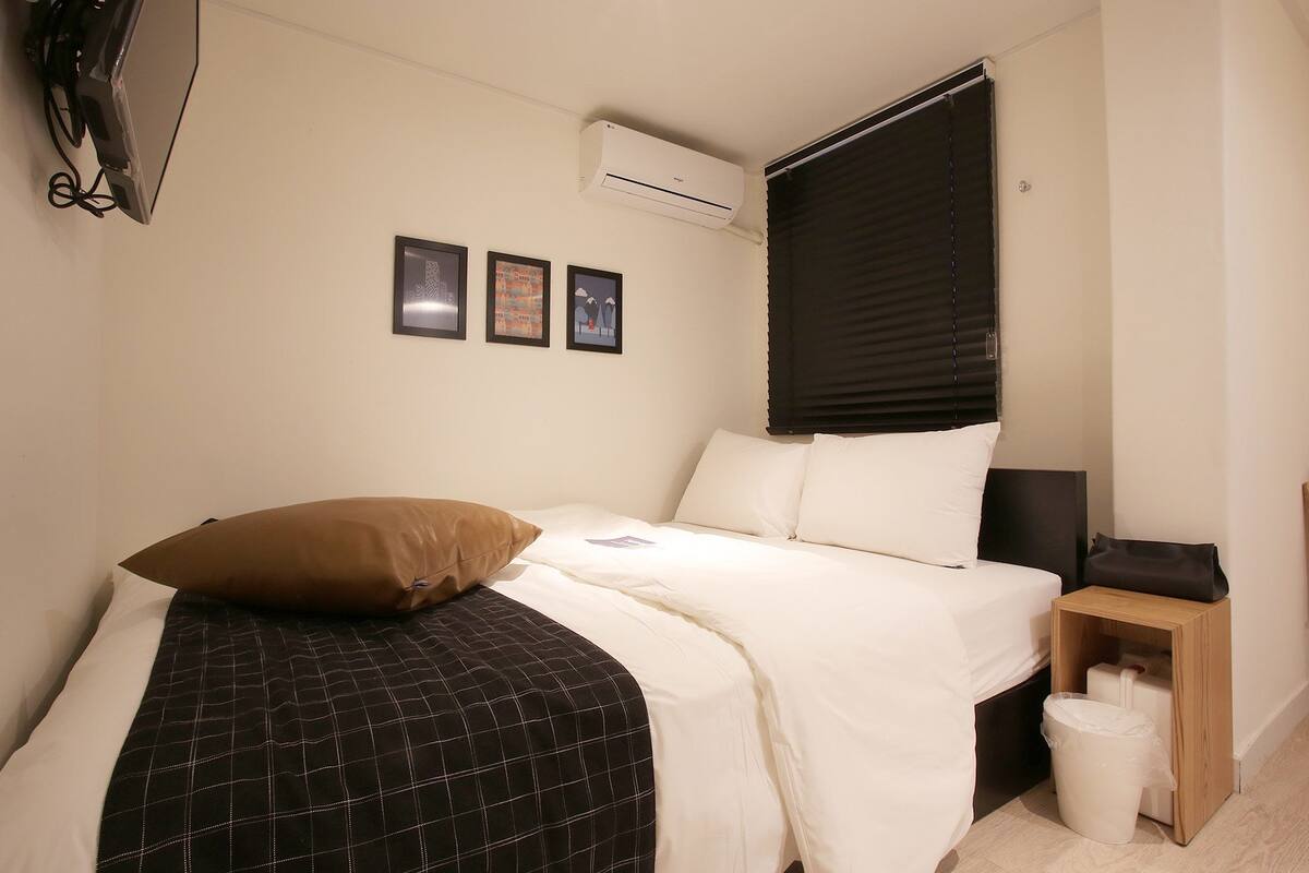 A neatly made bed with a brown pillow, a white pillow, and a black and white checkered blanket. There is a small wooden nightstand with a black bag and a white trash bin beside the bed.