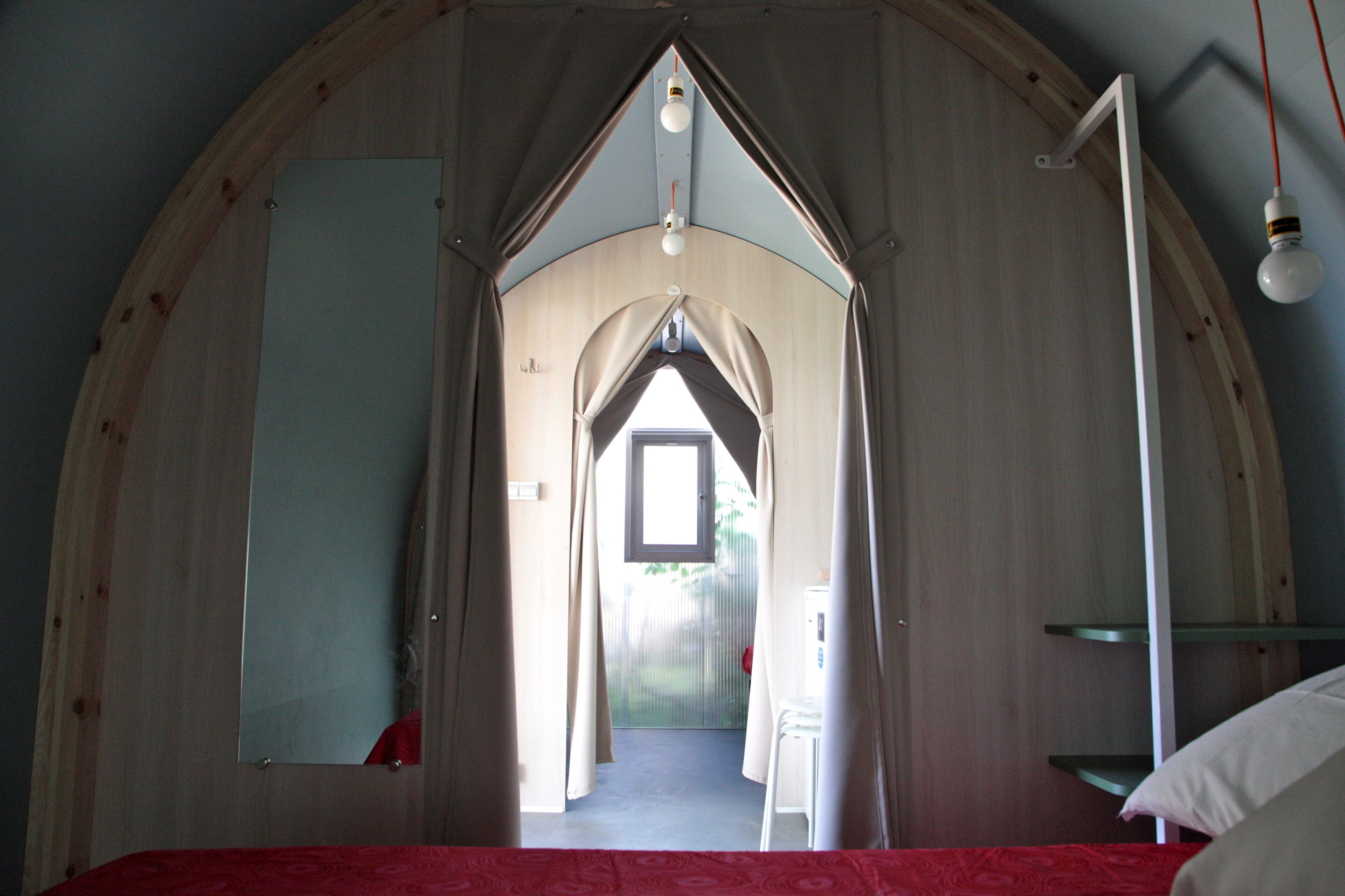 A room with a curved wooden ceiling, a bed with a red bedspread, a white bedside table, and a small window.
