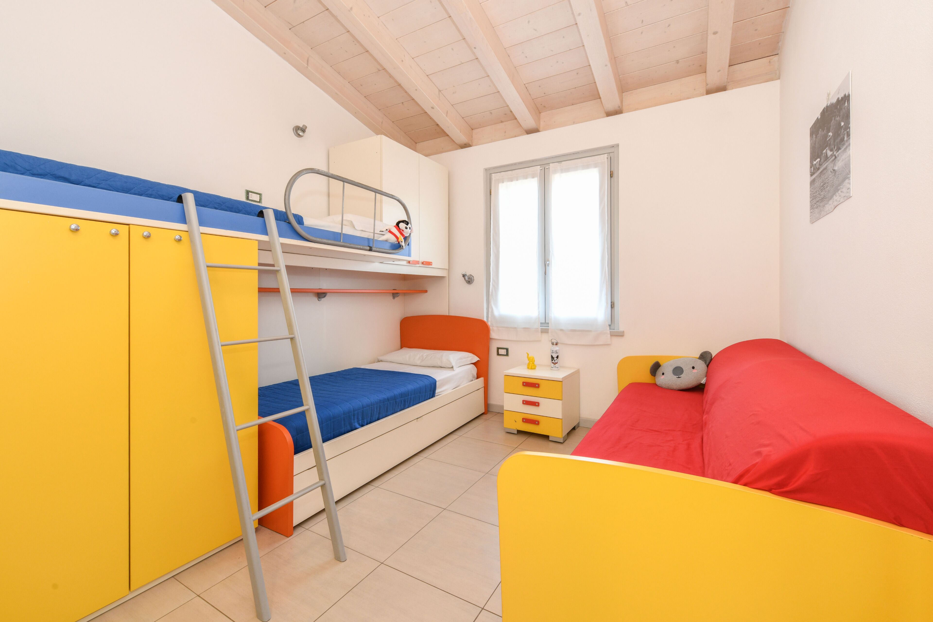 Une chambre avec des lits superposés, une échelle, une fenêtre et une petite table sur laquelle est posé un ours en peluche.