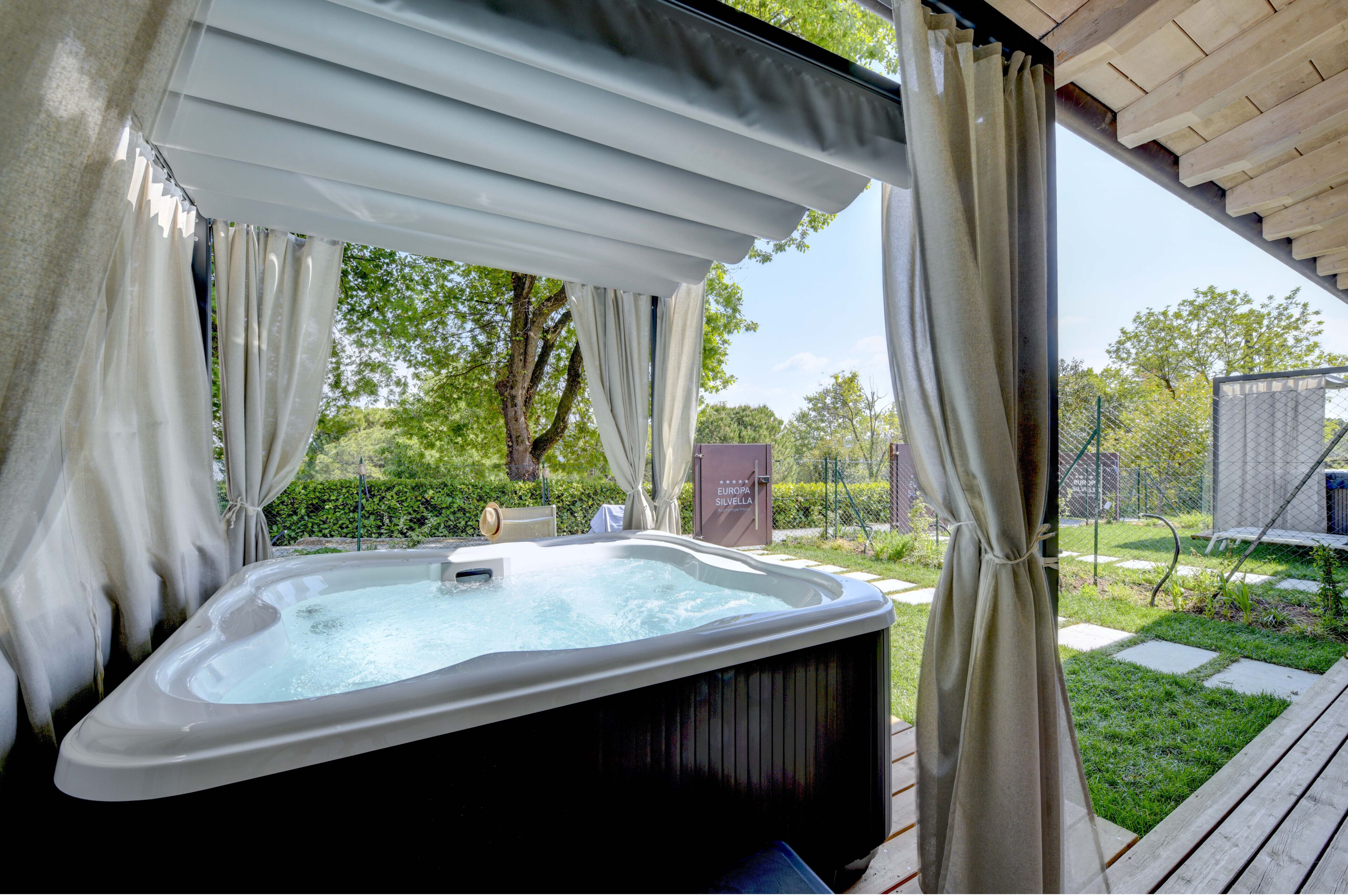 A hot tub under a pergola with curtains, surrounded by a garden and a fence.