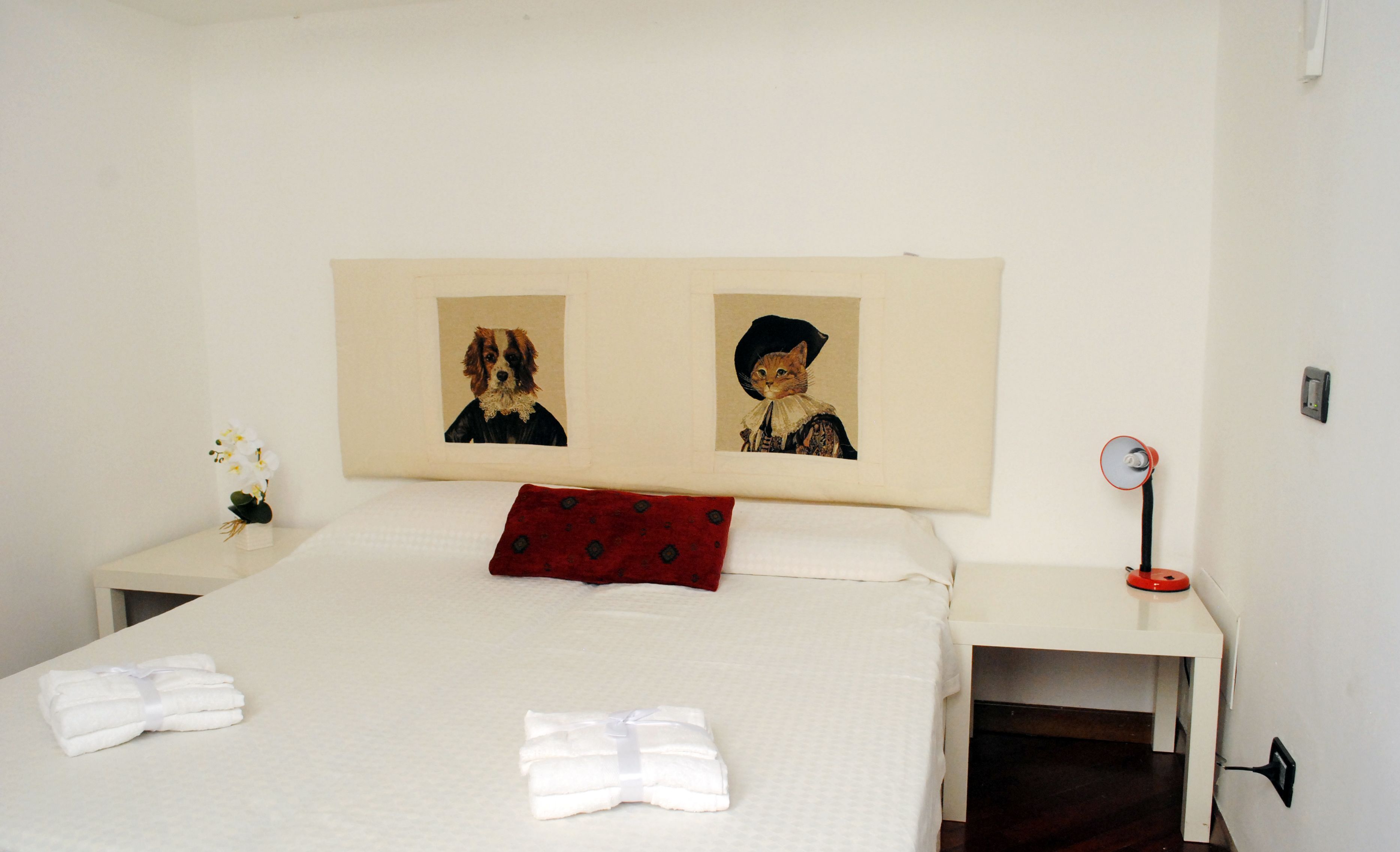 A neatly made bed with white linens and a red pillow, two framed pictures on the wall, a small vase with white flowers, and a red and black round mirror on a bedside table.