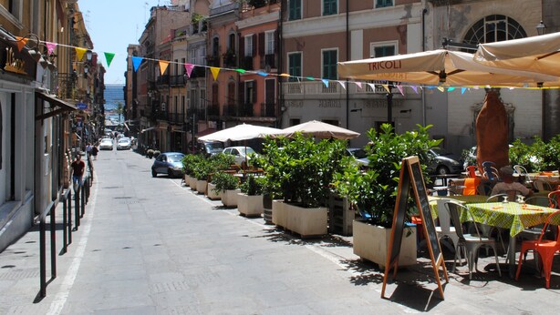 Una calle estrecha bordeada de edificios, con mesas al aire libre, sombrillas y plantas en macetas.