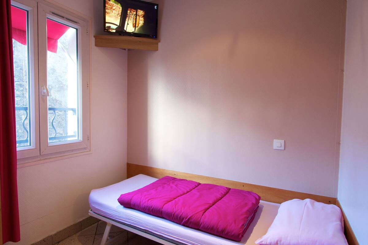 A small room with a single bed, a TV mounted on the wall, and a window with a red curtain.