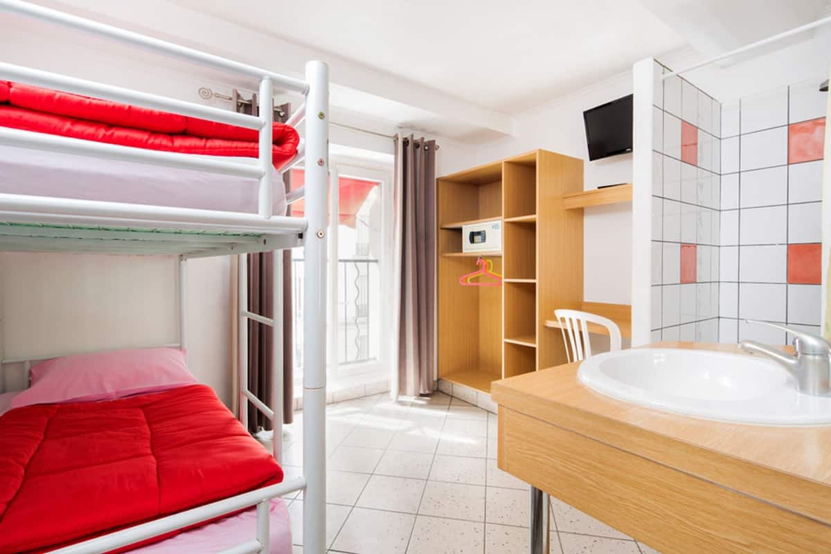 A bunk bed with red bedding, a wooden cabinet, a white chair, a sink, and a tiled wall.