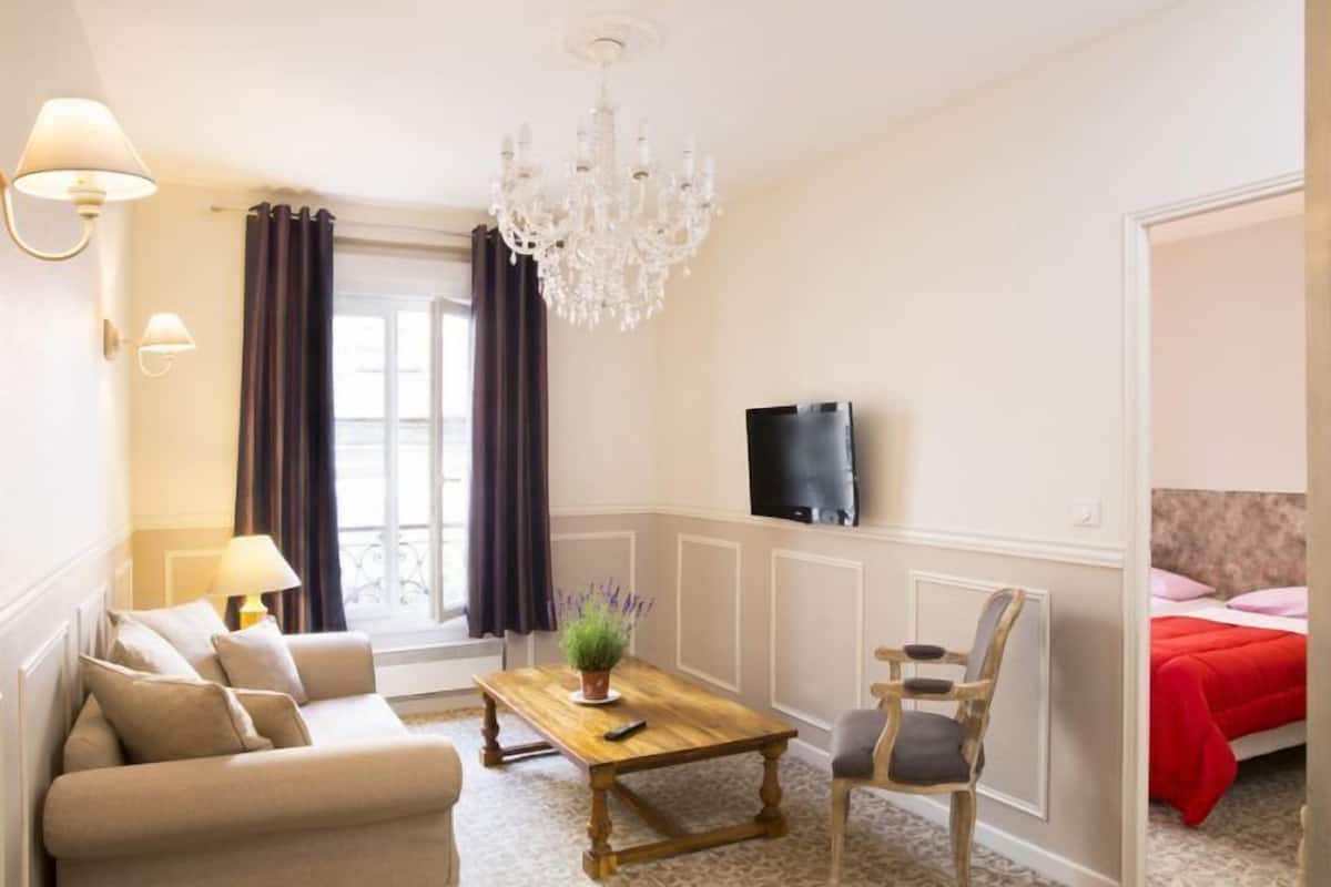 A hotel room with a wooden dining table, a sofa, a chair, a TV, and a chandelier.