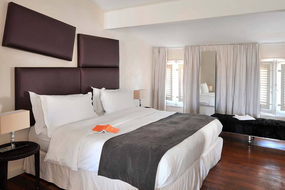 Habitación de hotel con una cama grande, una mesita de noche con lámpara, un espejo y vistas al exterior por la ventana.