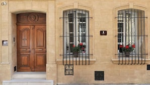 Un edificio con una amplia puerta doble de madera, dos ventanas arqueadas con rejas de metal y macetas con flores rojas en los alféizares.