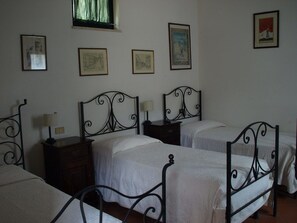 Un dormitorio con tres camas, cada una con estructura de metal y ropa de cama blanca. Hay cuadros enmarcados en la pared, una ventana con cortinas y una mesita de noche pequeña con una lámpara.