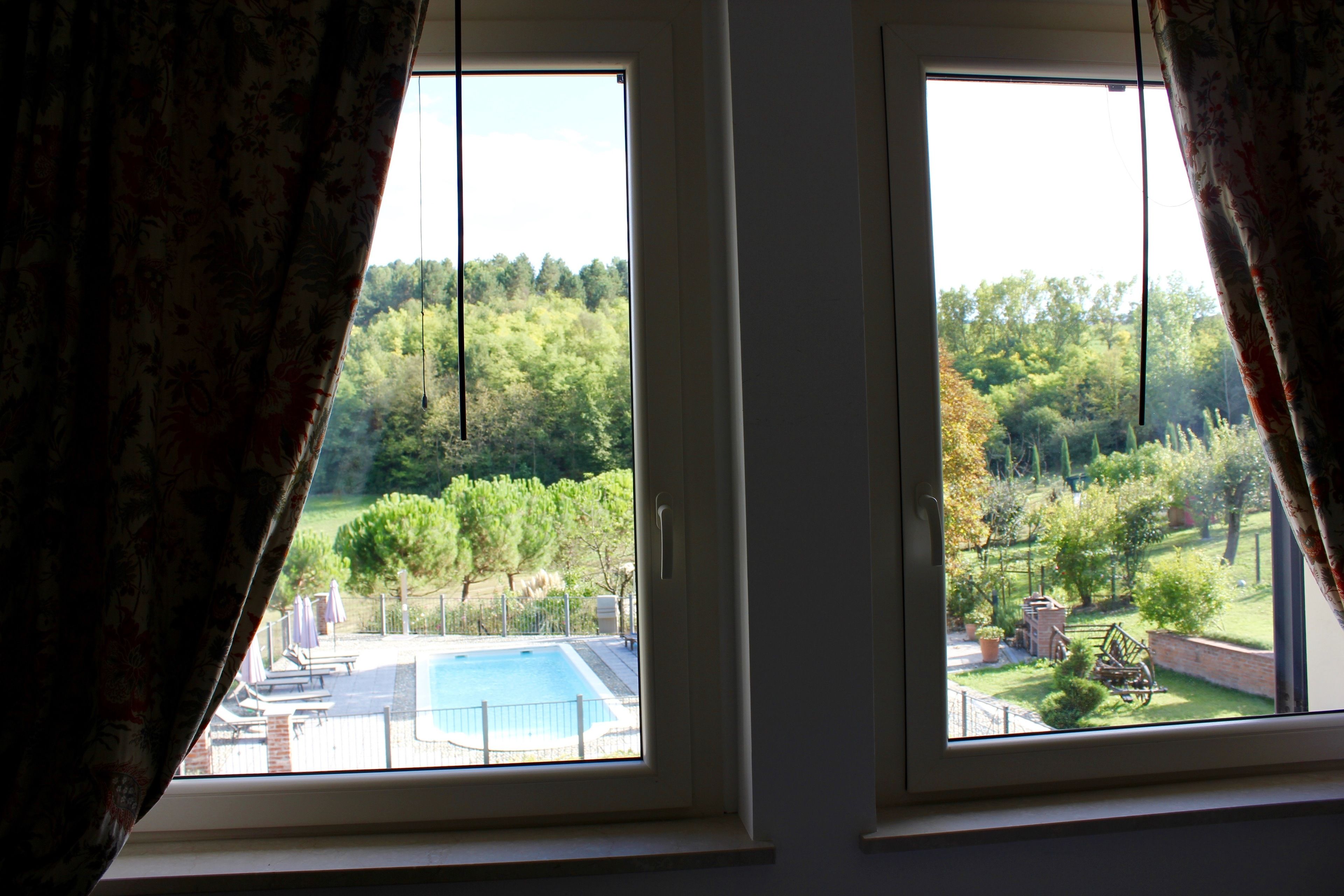 Dos ventanas con cortinas, vista a una piscina y un jardín con árboles y una estructura de ladrillo.