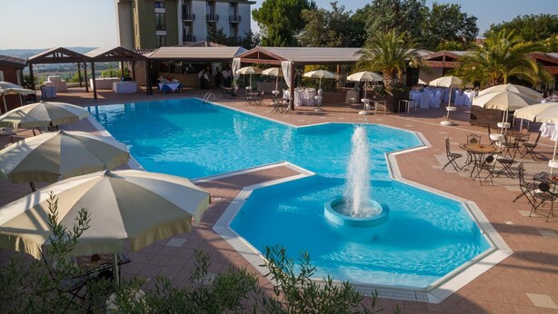 A swimming pool with a central fountain, surrounded by lounge chairs and umbrellas, with a building in the background. - La Cantinella (Ostra)