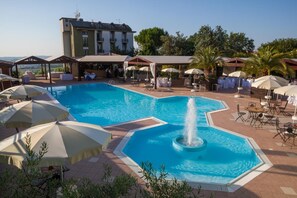 A swimming pool with a central fountain, surrounded by lounge chairs and umbrellas, with a building in the background. - La Cantinella (Ostra)