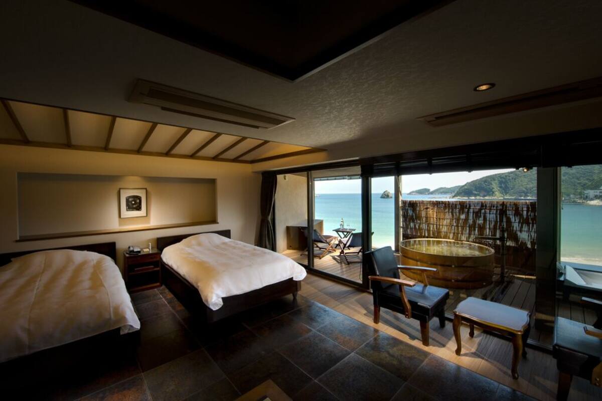 Une chambre d’hôtel avec deux lits, une grande fenêtre donnant sur la mer et une terrasse en bois avec un jacuzzi.