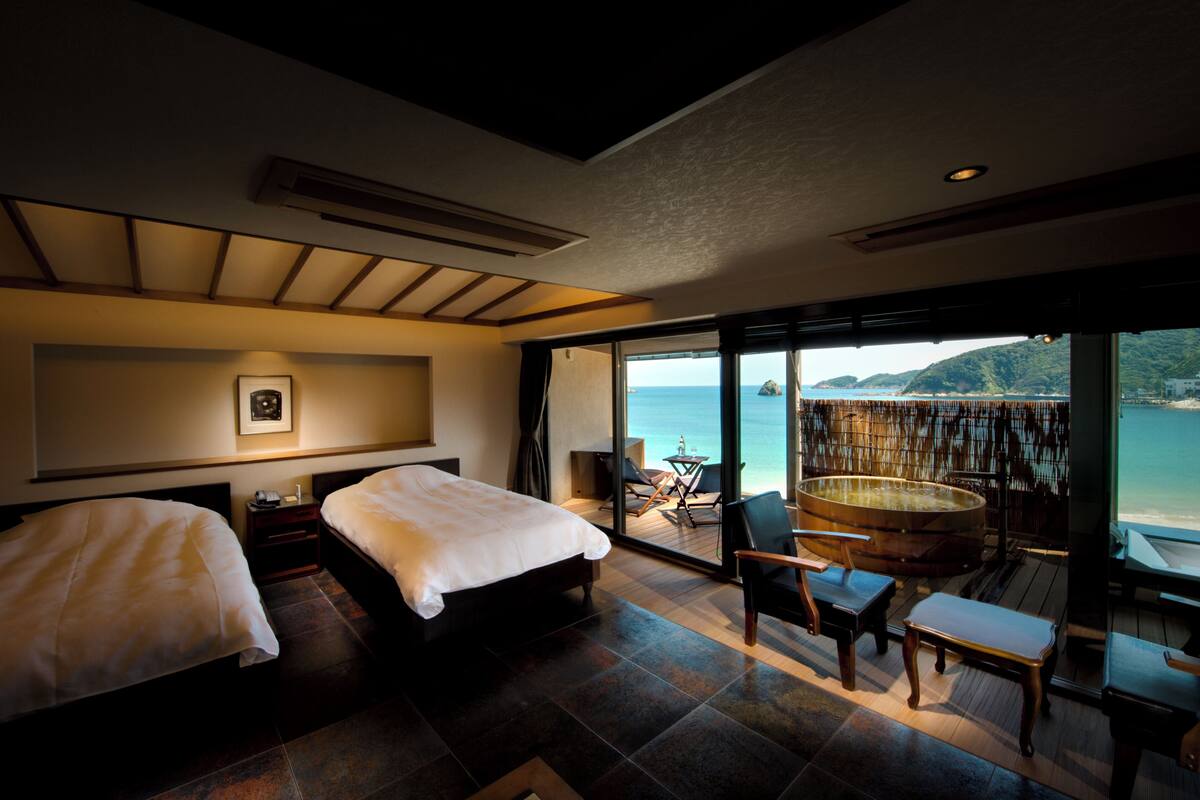 Une chambre d’hôtel avec deux lits, une grande fenêtre donnant sur la mer, une terrasse en bois et un jacuzzi.