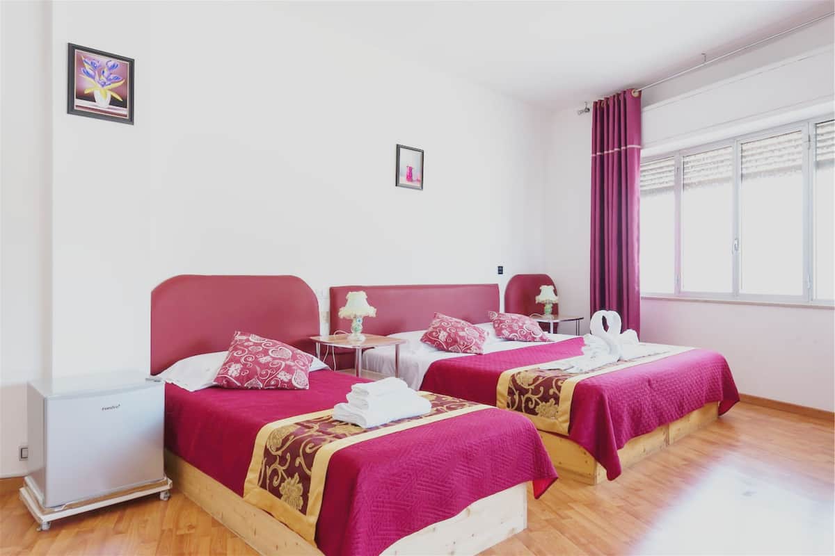 Habitación de hotel con dos camas, cada una con cabecera y colcha roja a juego. Cada cama tiene una mesita de noche con lámpara. La habitación tiene paredes blancas, una ventana con cortinas y un cuadro en la pared.