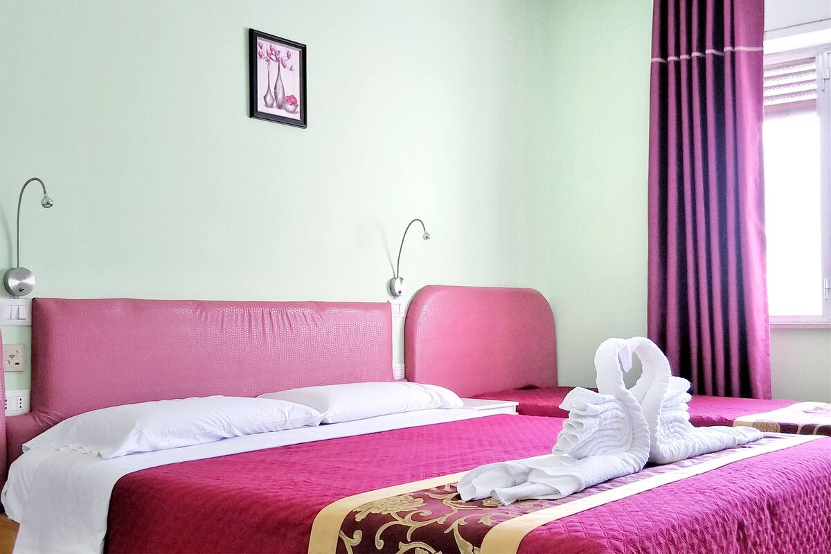 Habitación de hotel con dos camas, cada una con cabecera rosa y ropa de cama blanca. Hay un cuadro en la pared y una ventana con cortinas moradas.