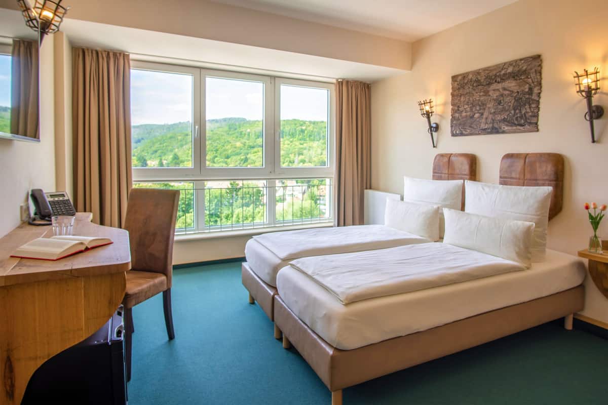 A hotel room with a large window, a desk with a telephone, a chair, a bed with pillows, and wall-mounted lamps.