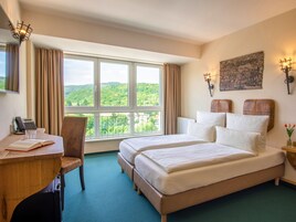 A hotel room with a large window, a desk with a telephone, a chair, a bed with pillows, and wall-mounted lamps.