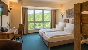 A hotel room with a large bed, a desk with a telephone, a chair, and a view of a green landscape through the window.