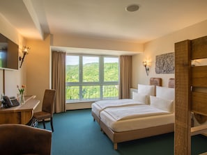 A hotel room with a large bed, a desk with a telephone, a chair, and a view of a green landscape through the window. - Hasseröder Burghotel (Wernigerode)