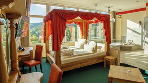A bedroom with a canopy bed, a desk, a chair, a small table, and a bathroom with a bathtub and a mirror. - Hasseröder Burghotel (Wernigerode)