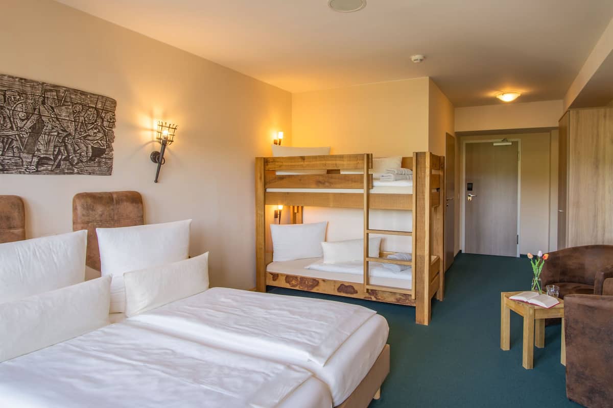 A hotel room with a large bed, a bunk bed, a small table with a book and flowers, and a door leading to another room.