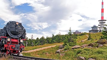 Una locomotora de vapor sobre rieles, con un paisaje montañoso y una alta torre roja y blanca al fondo.