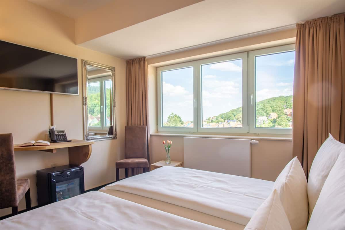 A hotel room with two beds, a desk with a telephone, a chair, a mirror, and a window with a view of greenery.
