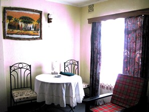 A room with a round table, two chairs, a painting on the wall, and a window with curtains.