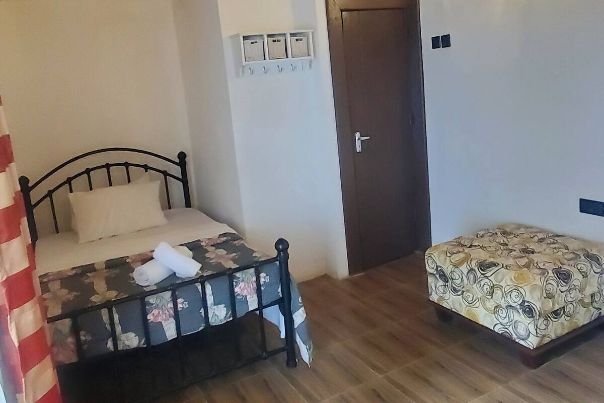 Quarto com cama, um banquinho, ventoinha no teto, porta e luminárias fixadas na parede.