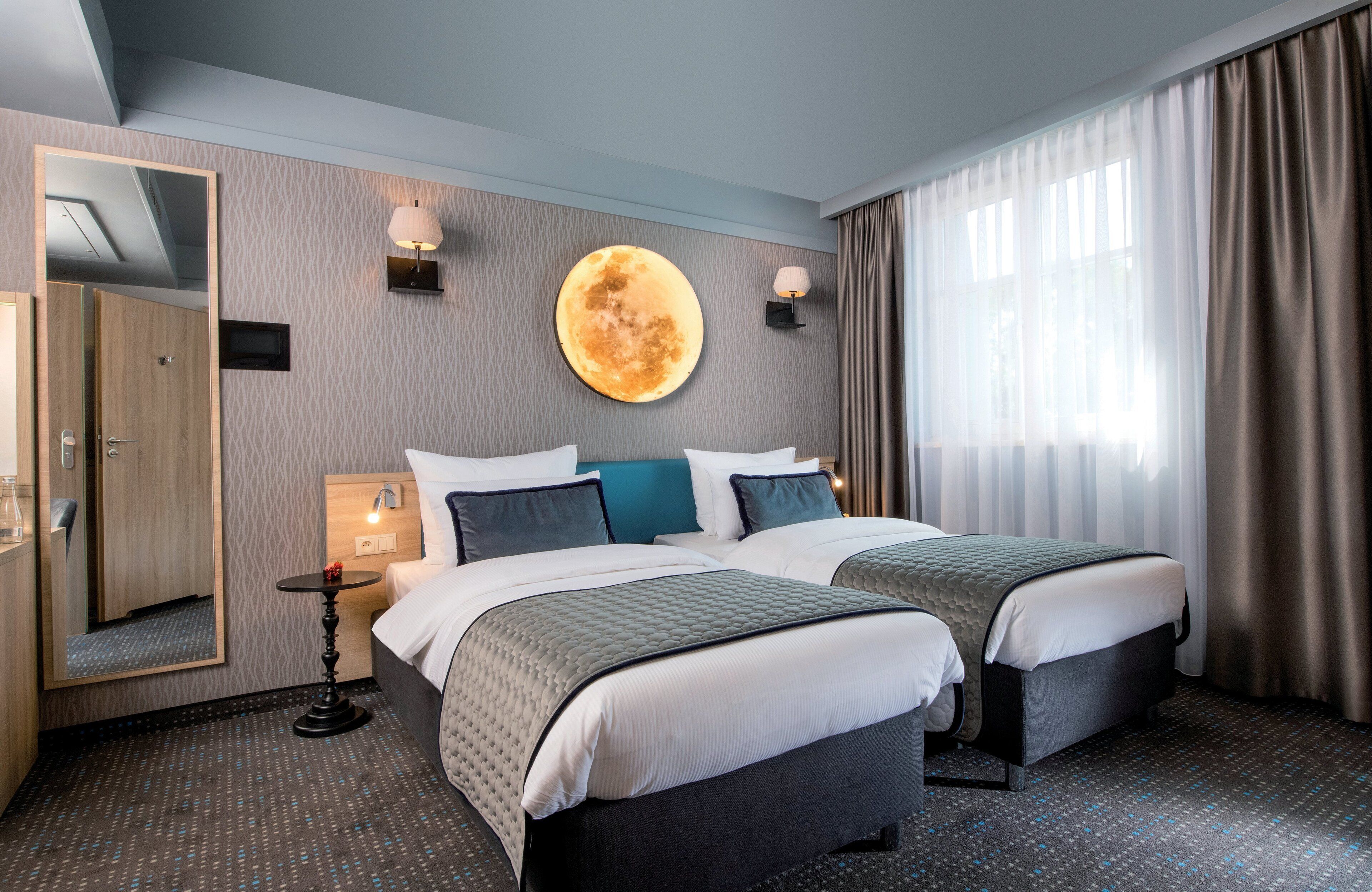 Ein Hotelzimmer mit zwei Betten, einem großen Spiegel, einer runden Wanddekoration und einem Fenster mit Vorhängen.
