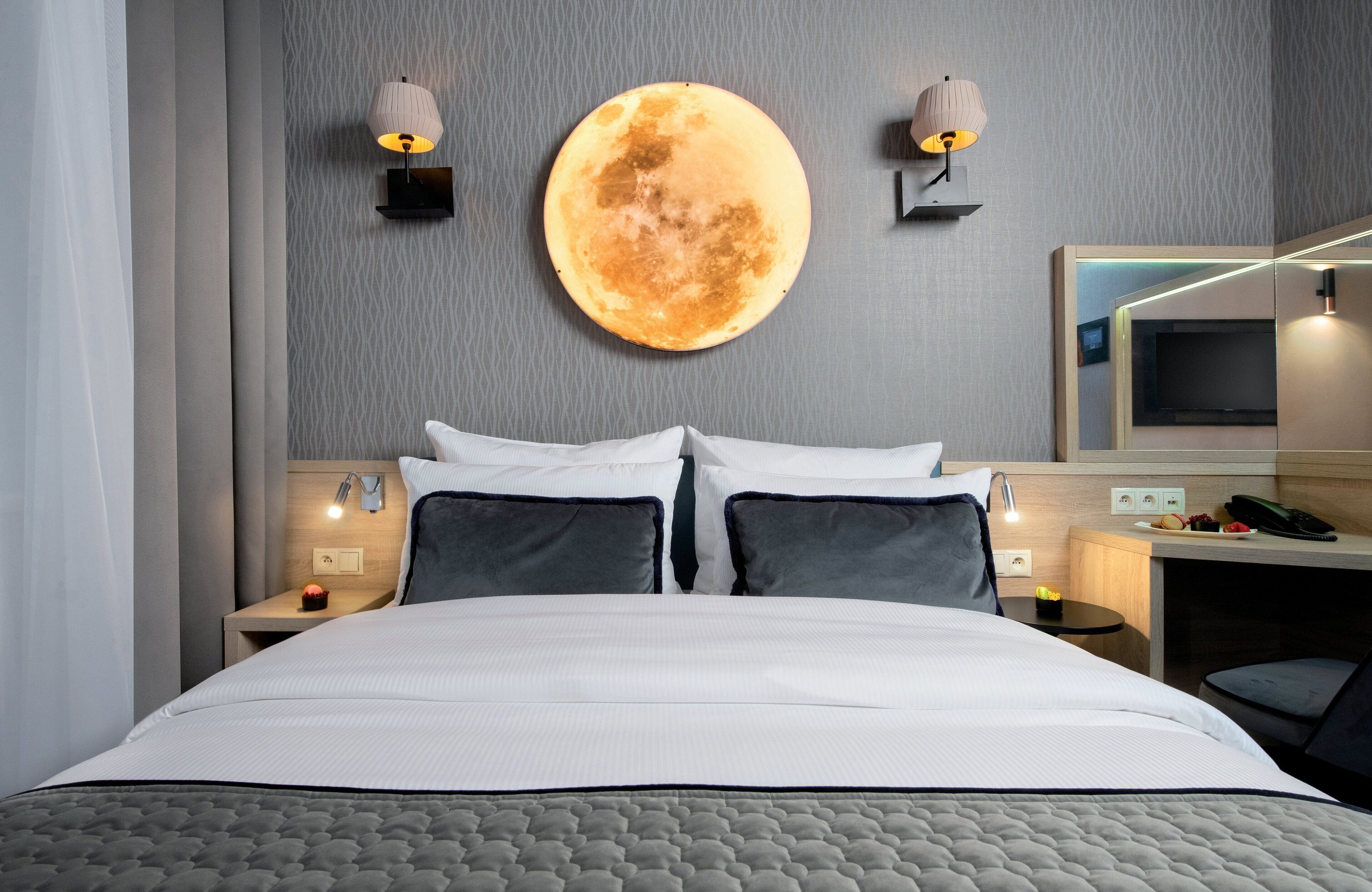A modern hotel room with a large bed, two bedside tables with lamps, a wall-mounted round mirror, and a flat-screen TV.