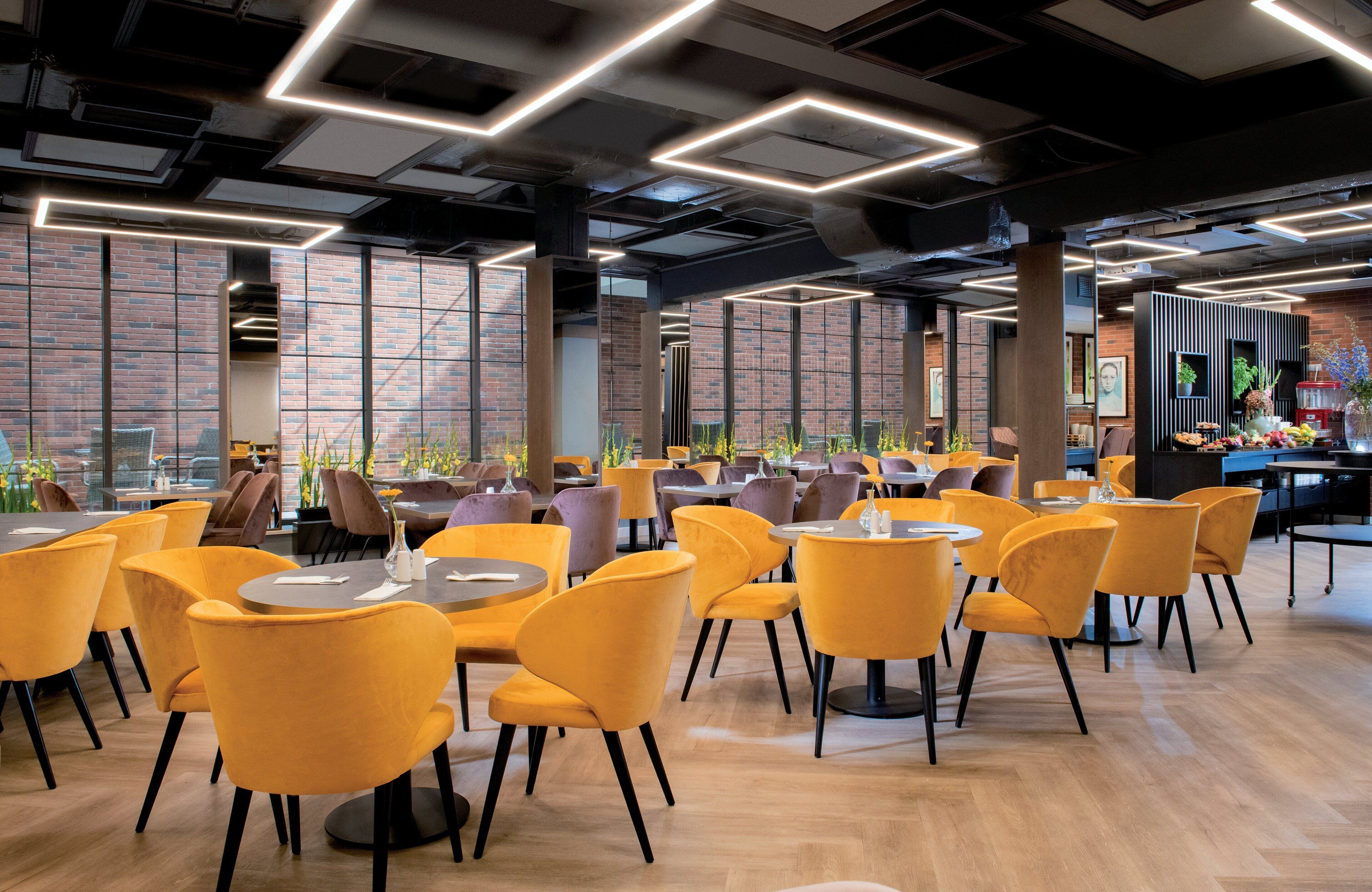 Un comedor moderno con sillas y mesas amarillas, paredes de ladrillo y ventanas grandes.