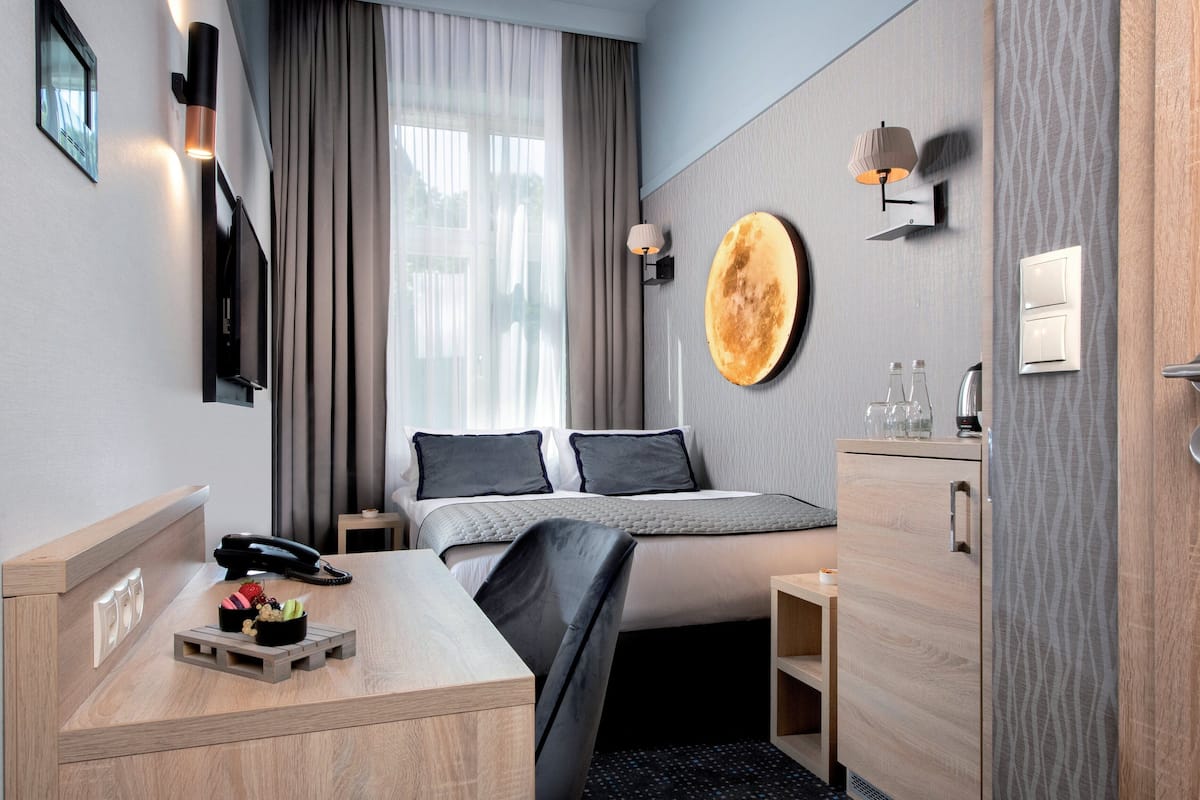 A modern hotel room with a bed, bedside table, lamp, and a wall-mounted round mirror.