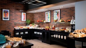A buffet setup with various food items and beverages, a coffee machine, and a brick wall with abstract art.