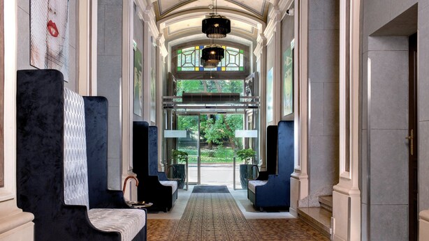 A grand hallway with a patterned carpet, seating areas with dark upholstery, and a view of greenery outside.