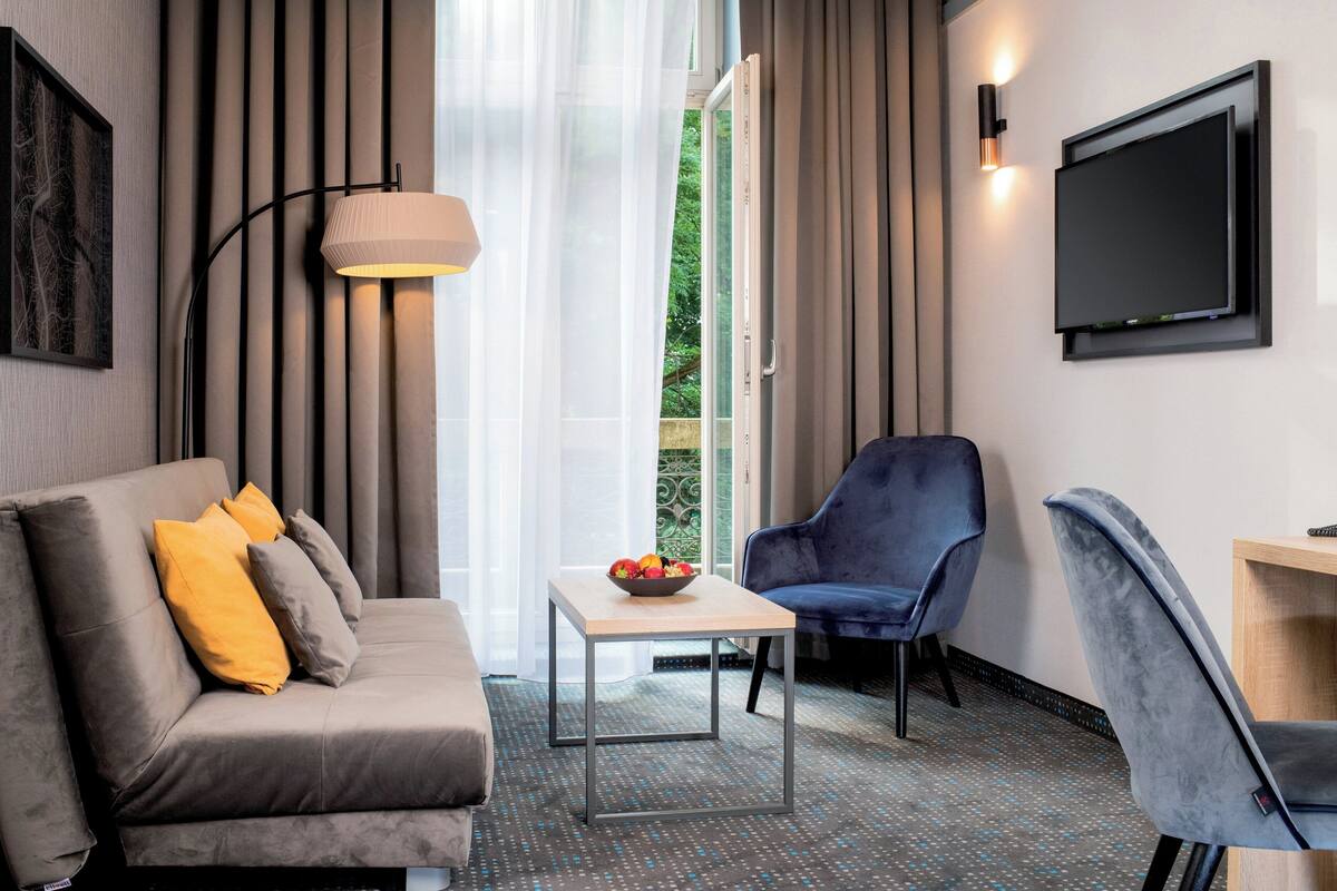 Une chambre d’hôtel moderne avec un canapé gris, un fauteuil bleu, une petite table sur laquelle se trouve un panier de fruits, un téléviseur fixé au mur et une fenêtre avec des rideaux.