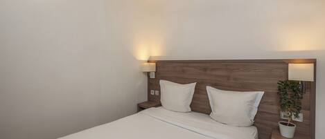 Una cama bien hecha con sábanas blancas, cabecera de madera, dos lámparas de noche y una pequeña planta en maceta sobre la mesita.