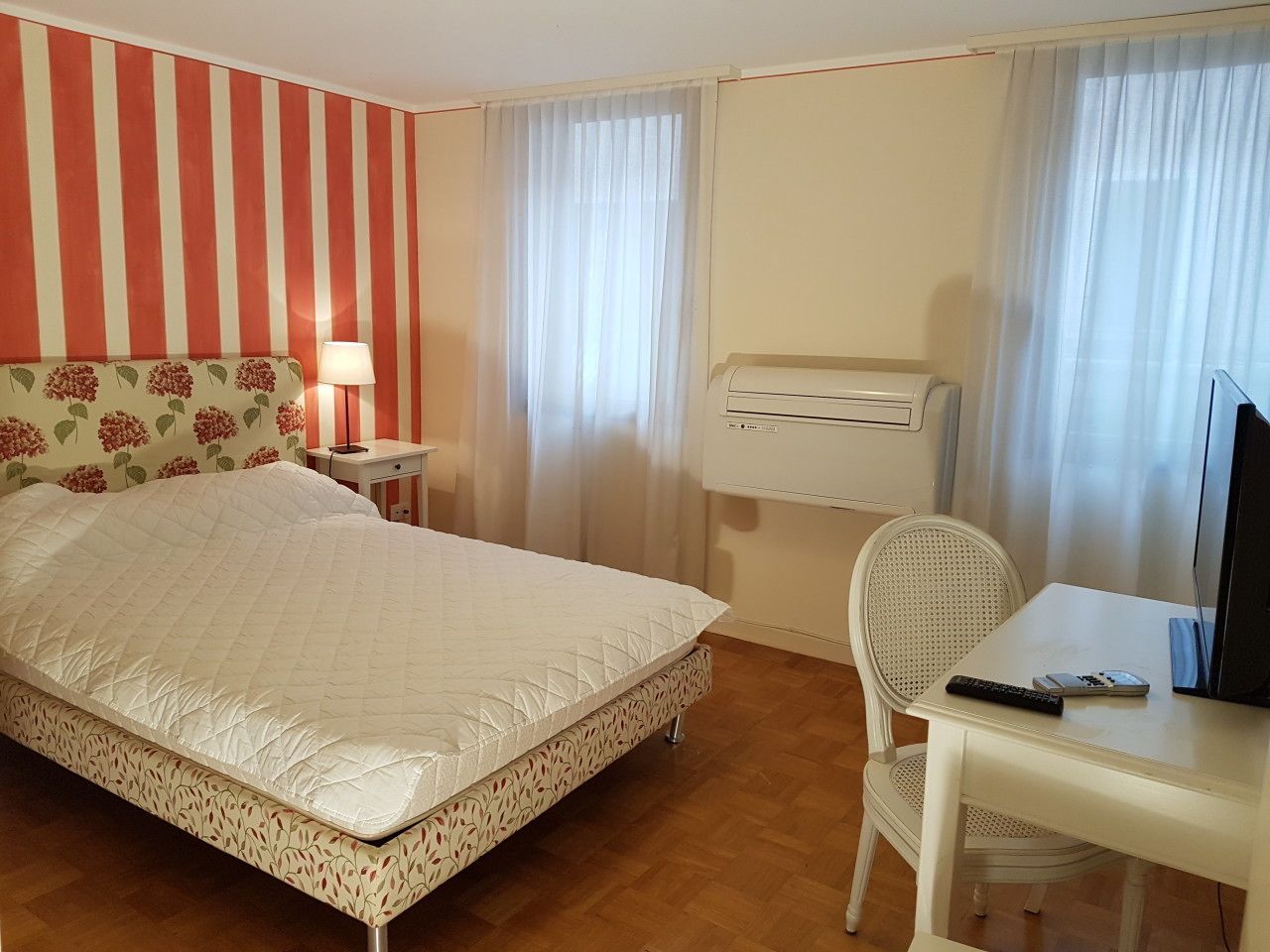 A hotel room with a bed, a desk with a computer, a chair, an air conditioner, and curtains.