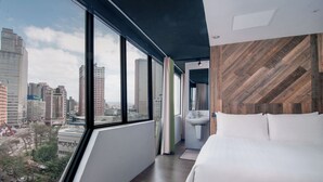 Habitación de hotel moderna con un ventanal amplio que ofrece vistas a la ciudad, una cama, un lavamanos y una pared de acento de madera.