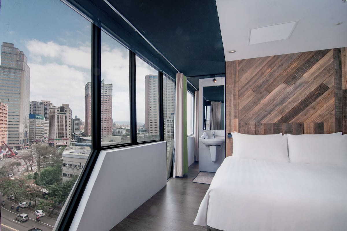 A modern hotel room with a large window offering a city view, a bed, a sink, and a wooden accent wall.