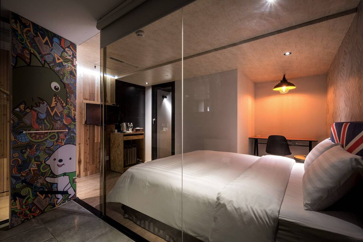 A modern hotel room with a large bed, a desk, and a chair. There is a glass partition separating the room from the hallway.