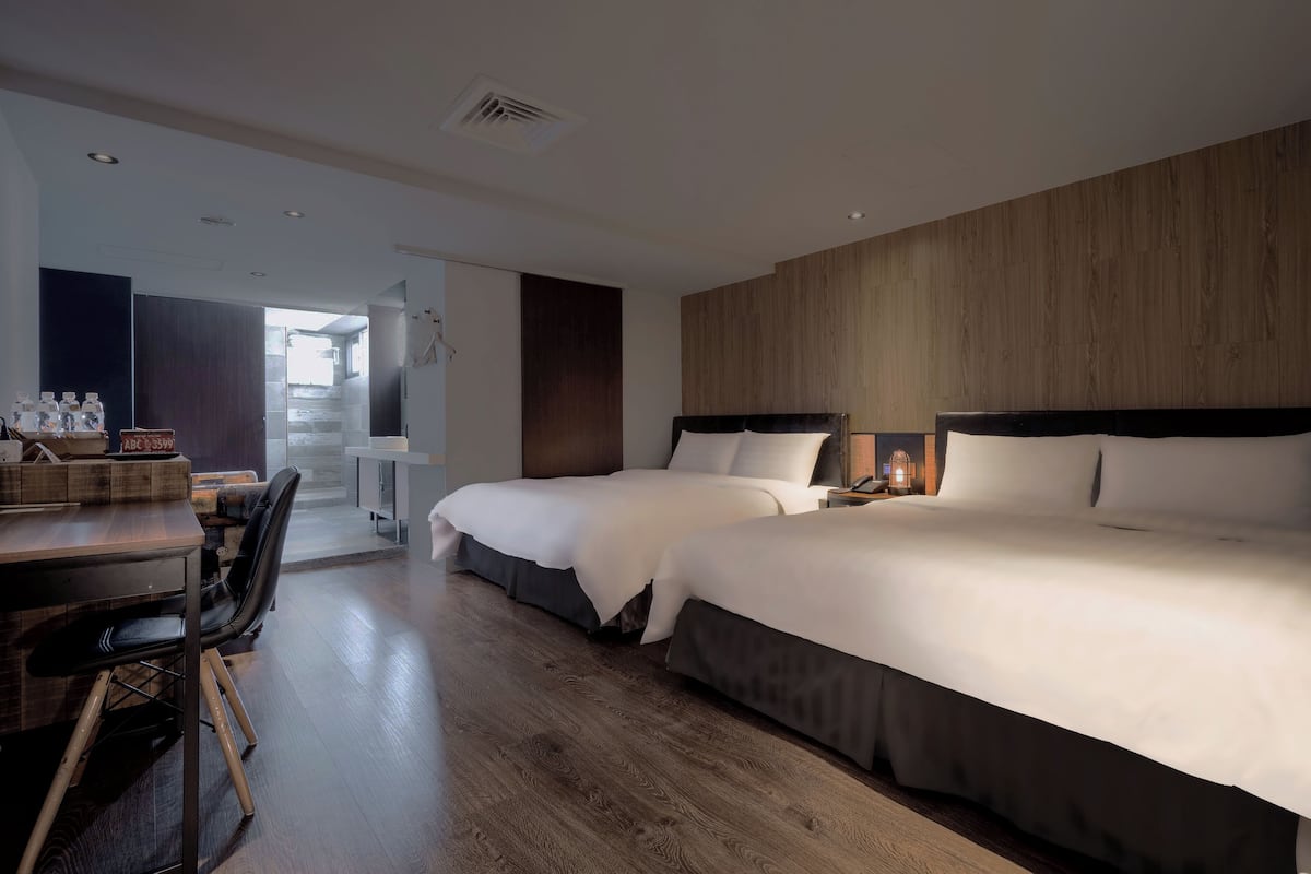 A hotel room with two beds, a wooden floor, a desk with chairs, and a view of the outside.