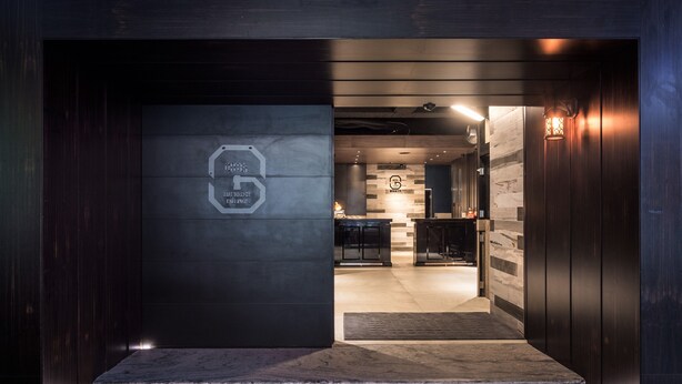 Entrada de un hotel moderno con fachada oscura, un logotipo geométrico en la pared y un interior bien iluminado que conduce a una zona de recepción.