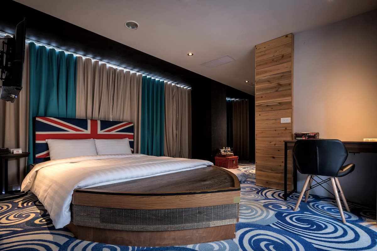 A modern hotel room with a large bed, a desk, and a chair. There is a British flag headboard and a red suitcase on the desk.