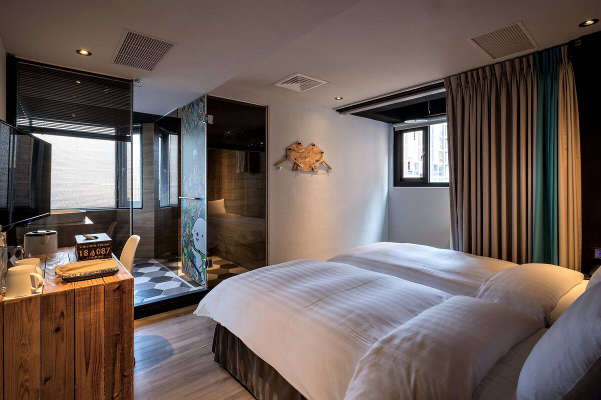 A modern hotel room with a large bed, a desk with a chair, a large window with curtains, and a wall-mounted artwork.