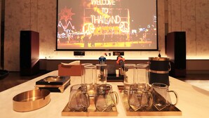 Zona de bar con una pantalla mostrando 'WELCOME TO THAILAND' y diversas herramientas de bar y vasos sobre la barra.