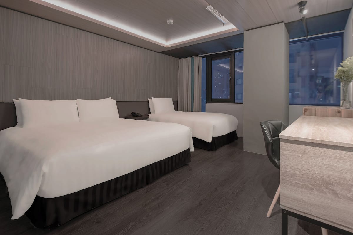 A modern hotel room with a large bed, a smaller bed, a desk, and a view of the city at night.